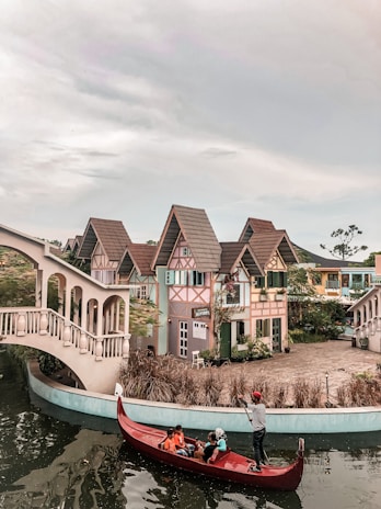 A picturesque scene featuring a quaint village with charming, colorful houses sporting steep, tiled roofs reminiscent of European architecture. A canal runs in the foreground, spanned by an elegant, arched bridge. A person navigates a red gondola on the water, carrying several passengers, adding a touch of Venetian charm to the setting. The area is lush with greenery, complementing the whimsical feel of the village.