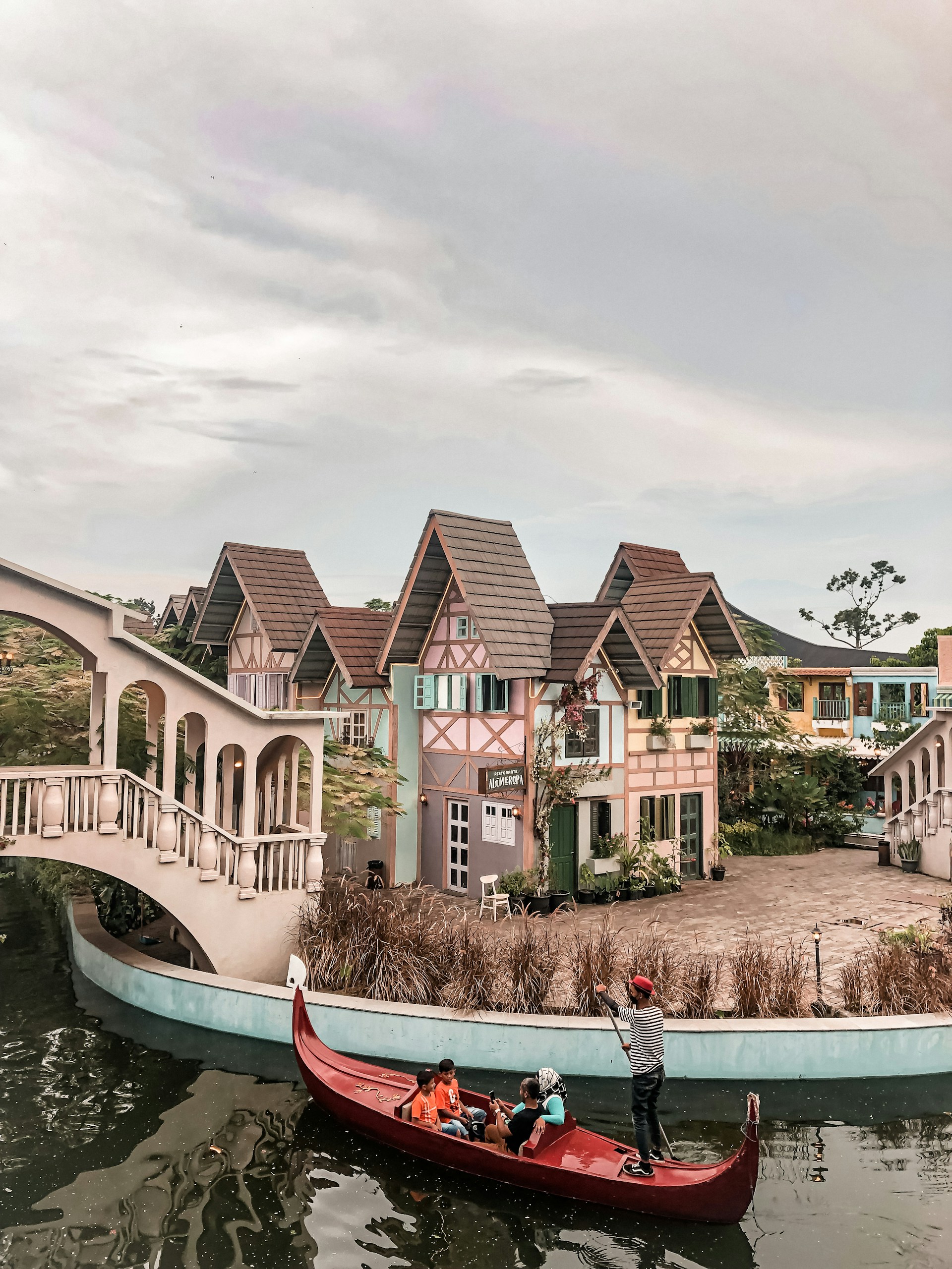 A picturesque scene featuring a quaint village with charming, colorful houses sporting steep, tiled roofs reminiscent of European architecture. A canal runs in the foreground, spanned by an elegant, arched bridge. A person navigates a red gondola on the water, carrying several passengers, adding a touch of Venetian charm to the setting. The area is lush with greenery, complementing the whimsical feel of the village.