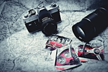 A vintage camera rests on a black and white map alongside photographs of a red sports car, featuring close-up shots of the car's interior and exterior. A large camera lens is positioned nearby, and the items are arranged in an artistic, nostalgic style.