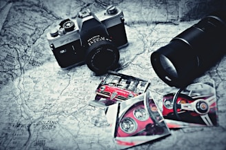 A vintage camera rests on a black and white map alongside photographs of a red sports car, featuring close-up shots of the car's interior and exterior. A large camera lens is positioned nearby, and the items are arranged in an artistic, nostalgic style.