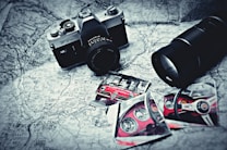 A vintage camera rests on a black and white map alongside photographs of a red sports car, featuring close-up shots of the car's interior and exterior. A large camera lens is positioned nearby, and the items are arranged in an artistic, nostalgic style.