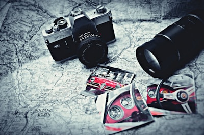 A vintage camera rests on a black and white map alongside photographs of a red sports car, featuring close-up shots of the car's interior and exterior. A large camera lens is positioned nearby, and the items are arranged in an artistic, nostalgic style.
