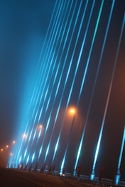 Tall structural cables of a modern bridge are dramatically illuminated with blue lights against a foggy background, with streetlights casting a warm yellow-orange glow.