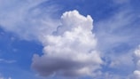 A large, fluffy cumulus cloud dominates the sky, with a backdrop of varying shades of blue and some wispy cirrus clouds. The cloud appears soft and billowy with a clean, white color contrasting against the bright blue sky.