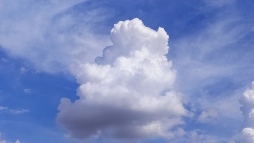 A large, fluffy cumulus cloud dominates the sky, with a backdrop of varying shades of blue and some wispy cirrus clouds. The cloud appears soft and billowy with a clean, white color contrasting against the bright blue sky.