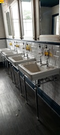 A row of three white ceramic sinks with metal faucets is mounted against a white tiled wall. Above them are mirrors and containers of liquid soap mounted on the wall. The floor is made of dark wood, adding contrast to the bright and clean aesthetic of the space.
