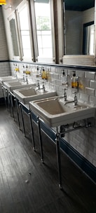 A row of three white ceramic sinks with metal faucets is mounted against a white tiled wall. Above them are mirrors and containers of liquid soap mounted on the wall. The floor is made of dark wood, adding contrast to the bright and clean aesthetic of the space.