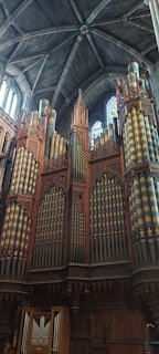 A grand and elaborate pipe organ positioned against a towering stone wall inside a cathedral. The organ features ornate wood carvings and multiple rows of pipes adorned with decorative patterns. Sunlight filters through a stained glass window above, casting colorful reflections onto the organ.