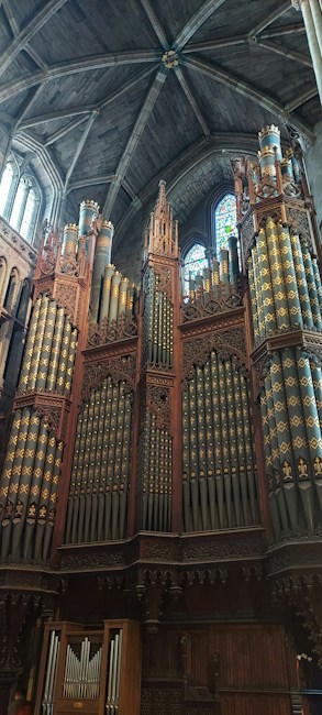 A grand and elaborate pipe organ positioned against a towering stone wall inside a cathedral. The organ features ornate wood carvings and multiple rows of pipes adorned with decorative patterns. Sunlight filters through a stained glass window above, casting colorful reflections onto the organ.