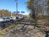 A busy roadway lined with cars in traffic, bordered by a footpath with one person walking away from the camera. The path is flanked by trees with sparse foliage, and there are various signs overhead.