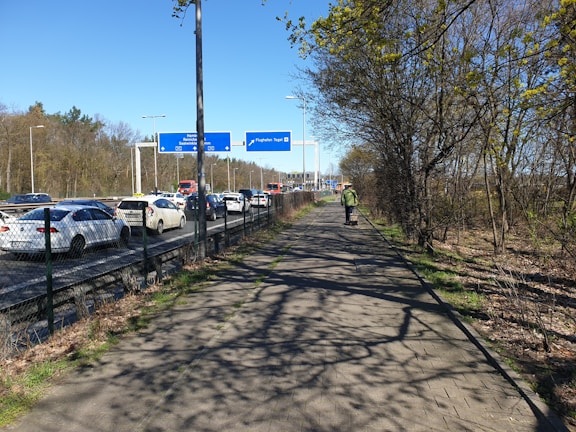 A busy roadway lined with cars in traffic, bordered by a footpath with one person walking away from the camera. The path is flanked by trees with sparse foliage, and there are various signs overhead.