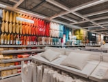 A retail store display featuring an extensive range of pillows arranged by color on shelves. The pillows vary from bright yellows, oranges, and reds to darker greens and blues. In the foreground, there are additional white pillows on display tables, with price tags visible. The setting appears organized and colorful, with fluorescent lighting from above.