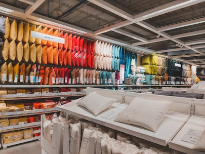 A retail store display featuring an extensive range of pillows arranged by color on shelves. The pillows vary from bright yellows, oranges, and reds to darker greens and blues. In the foreground, there are additional white pillows on display tables, with price tags visible. The setting appears organized and colorful, with fluorescent lighting from above.