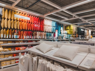 A retail store display featuring an extensive range of pillows arranged by color on shelves. The pillows vary from bright yellows, oranges, and reds to darker greens and blues. In the foreground, there are additional white pillows on display tables, with price tags visible. The setting appears organized and colorful, with fluorescent lighting from above.