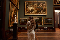 A visitor standing before a dense salon hang of framed paintings on a dark green wall