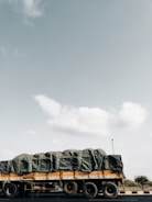 A large truck is seen on the road, carrying a heavily covered load with tarpaulin. The truck is yellow and occupies much of the lower portion of the scene. The sky is expansive and mostly clear, with a few scattered clouds visible.