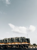 A large truck is seen on the road, carrying a heavily covered load with tarpaulin. The truck is yellow and occupies much of the lower portion of the scene. The sky is expansive and mostly clear, with a few scattered clouds visible.