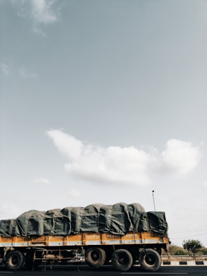 A large truck is seen on the road, carrying a heavily covered load with tarpaulin. The truck is yellow and occupies much of the lower portion of the scene. The sky is expansive and mostly clear, with a few scattered clouds visible.