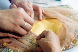 Hands are carefully sewing a piece of fabric, with one person holding a needle and thread while another adjusts delicate sheer material. The fabric beneath includes a mix of yellow and gold sequins, suggesting careful craftsmanship and attention to detail.