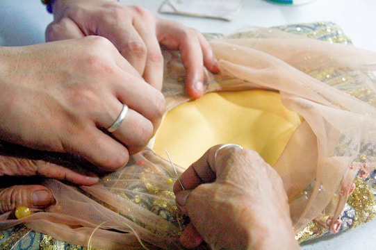 Hands are carefully sewing a piece of fabric, with one person holding a needle and thread while another adjusts delicate sheer material. The fabric beneath includes a mix of yellow and gold sequins, suggesting careful craftsmanship and attention to detail.