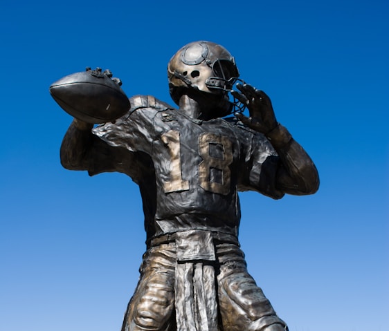 A bronze statue of an American football player in a dynamic pose, holding a football as if about to throw it. The player is wearing a helmet and jersey with the number 18. The sky in the background is clear and blue.