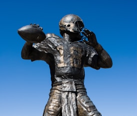 A bronze statue of an American football player in a dynamic pose, holding a football as if about to throw it. The player is wearing a helmet and jersey with the number 18. The sky in the background is clear and blue.
