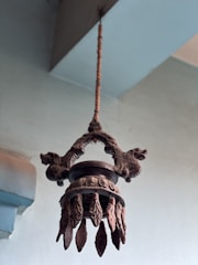 An intricately designed, antique metal chandelier hangs from the ceiling. It features ornate details and decorative elements, including several metal pieces resembling leaves or feathers hanging from the base. The background is a muted blue-green wall that complements the vintage aesthetic.