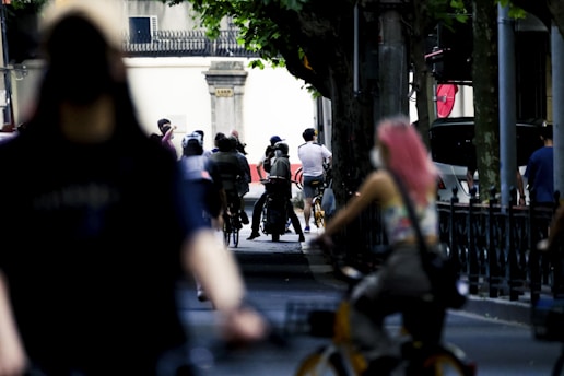 A city street scene features several people riding bicycles along a shaded path. The background shows a variety of blurred figures, street poles, and parked cars under a canopy of trees, creating a dynamic urban environment.