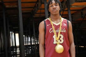 A young man is wearing a basketball jersey with the number 13 and the word 'Creekside' printed on it. He is also adorned with a large, gold chain that has a pendant shaped like a basketball. The background consists of dark, metal structures, possibly the underside of bleachers or seats.