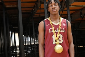 A young man is wearing a basketball jersey with the number 13 and the word 'Creekside' printed on it. He is also adorned with a large, gold chain that has a pendant shaped like a basketball. The background consists of dark, metal structures, possibly the underside of bleachers or seats.