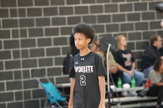 A young basketball player with an afro hairstyle stands in a gymnasium, wearing a black jersey with the number 2 and the text 'P3 ELITE'. Behind the player, there are spectators sitting on bleachers with a brick wall in the background.