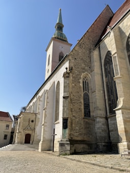 A tall, historic church with a pointed green steeple and a clock faces a cobblestone courtyard. The building features large arched windows and a mix of stone and plaster exterior, showing signs of age and architectural detail.