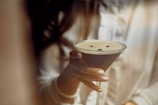 A hand with neatly manicured nails holds a martini glass filled with a dark, frothy cocktail, topped with coffee beans. The person is dressed in a cream-colored blouse with a pearl bracelet visible on the wrist.