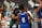 A young athlete wearing a blue jersey with the number 11 stands on a basketball court. Other players and people are visible in the background, including a player in a grey jersey. The setting appears to be a crowded indoor sports venue.