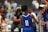 A young athlete wearing a blue jersey with the number 11 stands on a basketball court. Other players and people are visible in the background, including a player in a grey jersey. The setting appears to be a crowded indoor sports venue.