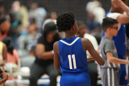 A young athlete wearing a blue jersey with the number 11 stands on a basketball court. Other players and people are visible in the background, including a player in a grey jersey. The setting appears to be a crowded indoor sports venue.