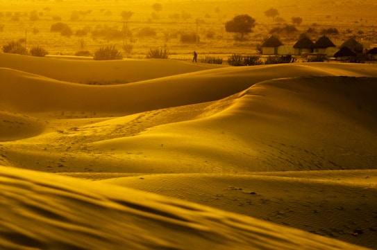 Golden sand dunes illuminated by warm sunset light, with shadows accentuating the curves. In the background, a silhouette of a person is walking near small huts and scattered bushes, creating a tranquil desert scene.