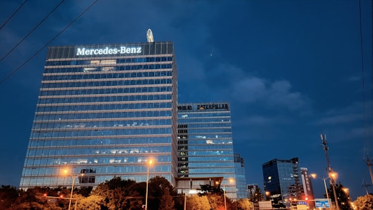 Tall office buildings with illuminated signs are seen against a twilight sky. The largest building prominently displays the Mercedes-Benz logo. The surrounding area is bathed in a warm glow from streetlights and some green foliage is visible at the bottom.