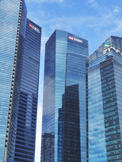 Tall modern skyscrapers with reflective glass facades dominate the scene, set against a clear blue sky. Each building features logos of various financial institutions.