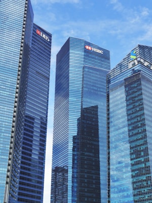 Tall modern skyscrapers with reflective glass facades dominate the scene, set against a clear blue sky. Each building features logos of various financial institutions.