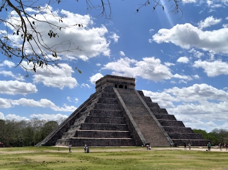 A large, ancient stone pyramid stands majestically against a backdrop of a bright blue sky with scattered white clouds. Surrounding the pyramid are open grassy areas and a few people can be seen walking nearby. Leafy branches are visible in the upper part of the image, adding a natural frame to the scene.