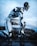 A humanoid robot with a sleek, futuristic design is kneeling on a rocky surface near a body of water. The robot's structure is composed of white and black components, showcasing exposed mechanical joints and complex circuitry. The sky above is overcast, contributing to the dramatic setting.