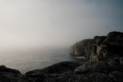 A mist-covered coastal landscape with rugged cliffs jutting over the ocean. The horizon is obscured by dense fog, blending into the sky, creating a mysterious and serene atmosphere. A lighthouse is barely visible in the distance on the headland.