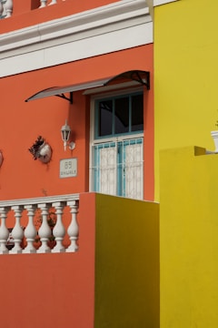 An architectural facade featuring vibrant orange and yellow walls with white decorative elements such as a balustrade and window frames. A small, stylized black metal awning hangs above a window, and a wall-mounted lamp and two ceramic decorations accent the wall. The area is well-lit, revealing intricate details.