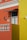 An architectural facade featuring vibrant orange and yellow walls with white decorative elements such as a balustrade and window frames. A small, stylized black metal awning hangs above a window, and a wall-mounted lamp and two ceramic decorations accent the wall. The area is well-lit, revealing intricate details.