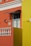 An architectural facade featuring vibrant orange and yellow walls with white decorative elements such as a balustrade and window frames. A small, stylized black metal awning hangs above a window, and a wall-mounted lamp and two ceramic decorations accent the wall. The area is well-lit, revealing intricate details.