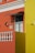 An architectural facade featuring vibrant orange and yellow walls with white decorative elements such as a balustrade and window frames. A small, stylized black metal awning hangs above a window, and a wall-mounted lamp and two ceramic decorations accent the wall. The area is well-lit, revealing intricate details.