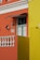 An architectural facade featuring vibrant orange and yellow walls with white decorative elements such as a balustrade and window frames. A small, stylized black metal awning hangs above a window, and a wall-mounted lamp and two ceramic decorations accent the wall. The area is well-lit, revealing intricate details.