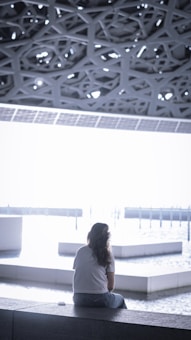 A person with long hair sits alone on a concrete bench, facing a bright waterfront. Above, a geometric ceiling design creates a patterned canopy. The atmosphere is peaceful and introspective.