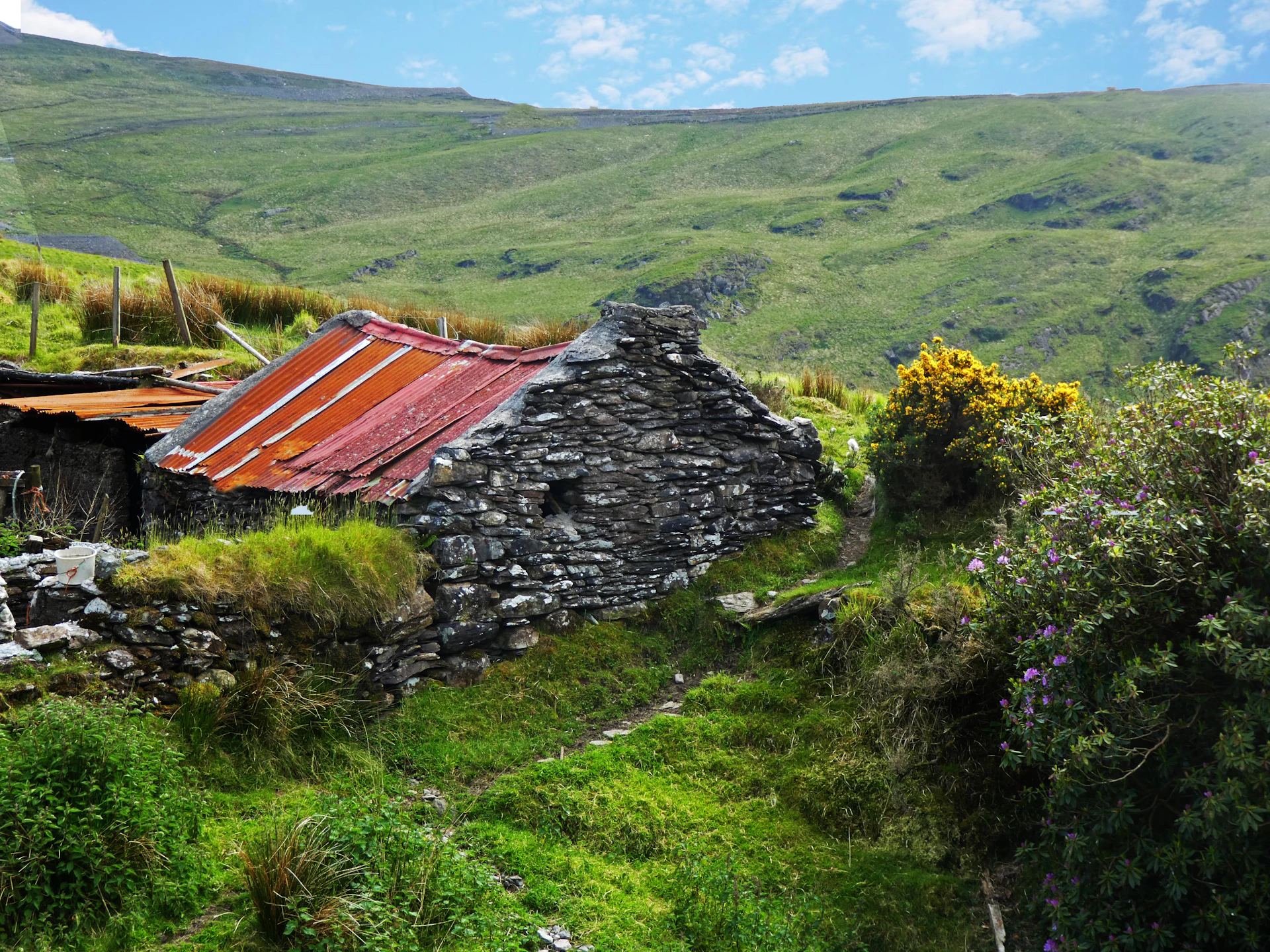 The rustic stone exterior of Le Roc Del Four cottage under a bright blue sky, with wildflowers blooming nearby.